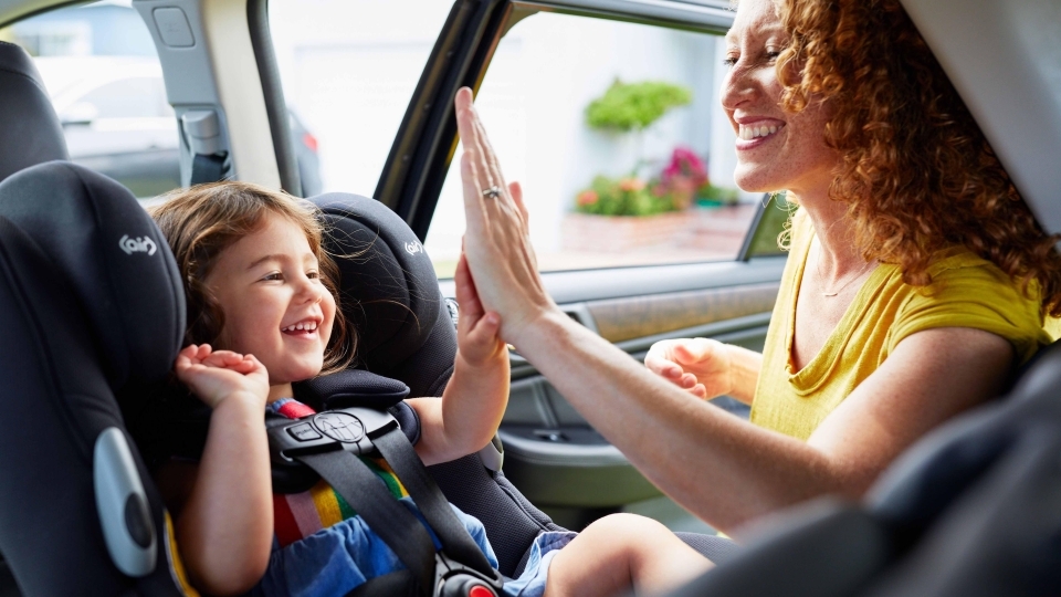 A smiling child sitting in a car seat high-fiving their mom